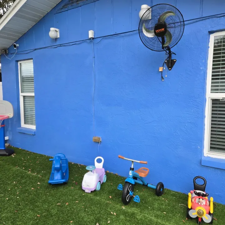 Outdoor play area with a tricycle, ride-on toys, and a basketball hoop against the daycare's exterior wall